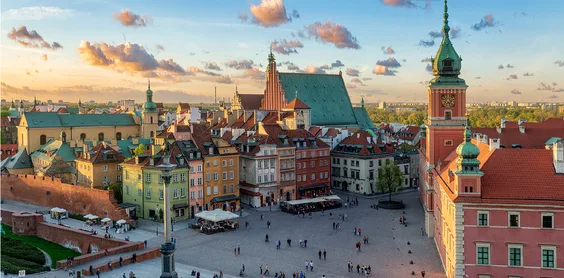View of Castle Square in Warsaw, Poland, with colorful buildings, a clock tower, a cathedral roof, and people walking.